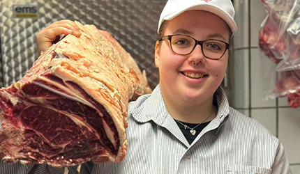 Fleischermeisterin hält einen frischen Strang Rinderrücken (Roastbeef) im Kühlhaus und lächelt stolz in die Kamera.