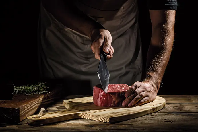 Fleischer bearbeitet ein rohes Stück Fleisch auf einem Holzbrett mit einem Beil.