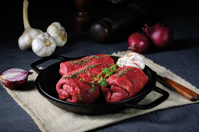Frische Rinderrouladen in einer gusseisernen Pfanne, umgeben von Knoblauch und Zwiebeln.