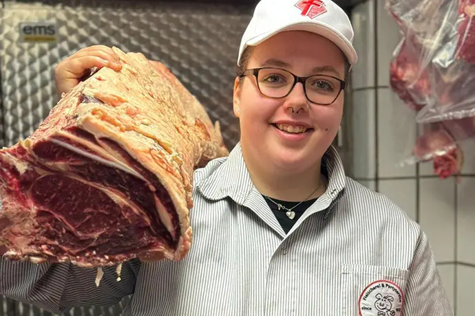 Fleischermeisterin hält einen frischen Strang Rinderrücken (Roastbeef) im Kühlhaus und lächelt stolz in die Kamera.