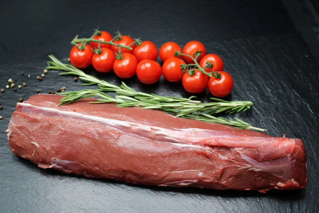 Roher Rehrückenlachs auf einer Schieferplatte, umgeben von Pfefferkörnern, Rosmarin und Tomaten.
